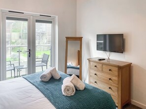 Quarto de hotel com uma cama, uma TV sobre uma cômoda de madeira e uma porta de vidro corrediça que dá para uma área externa com uma cadeira e mesa.