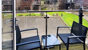 A balcony with two black chairs and a small table, overlooking a grassy area and buildings in the background.