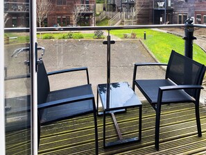 A balcony with two black chairs and a small table, overlooking a grassy area and buildings in the background.