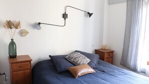 A bedroom with a bed, a wall-mounted lamp, a nightstand, a dresser, and a window with curtains. - Mas Provençal Rénové Luberon (La Bastide-des-Jourdans)