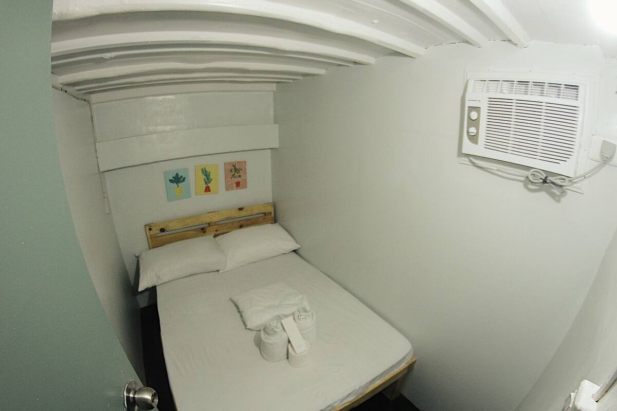 A small, single-bed room with a wooden headboard, a wall-mounted air conditioning unit, and three small potted plants on the wall.
