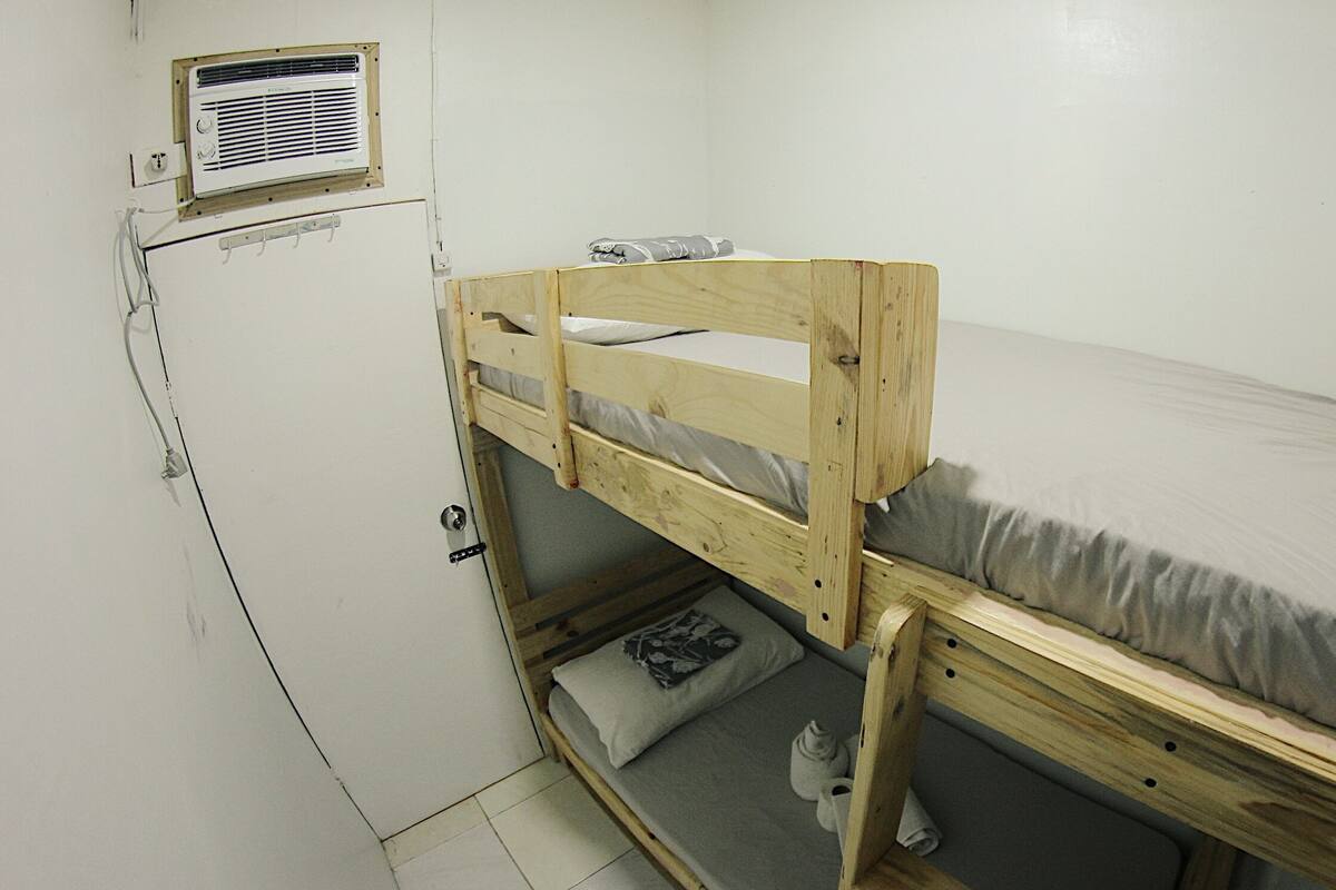 A small, single-bedded room with a wooden bunk bed, a small heater, and a wall-mounted air conditioning unit.
