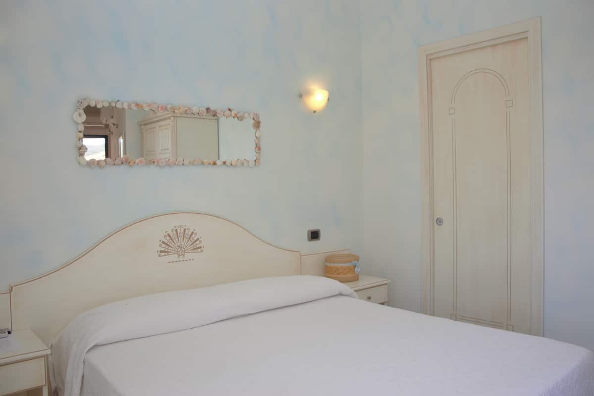 A bedroom with a bed, a nightstand, a mirror with a seashell frame, a wall-mounted light, and a door.
