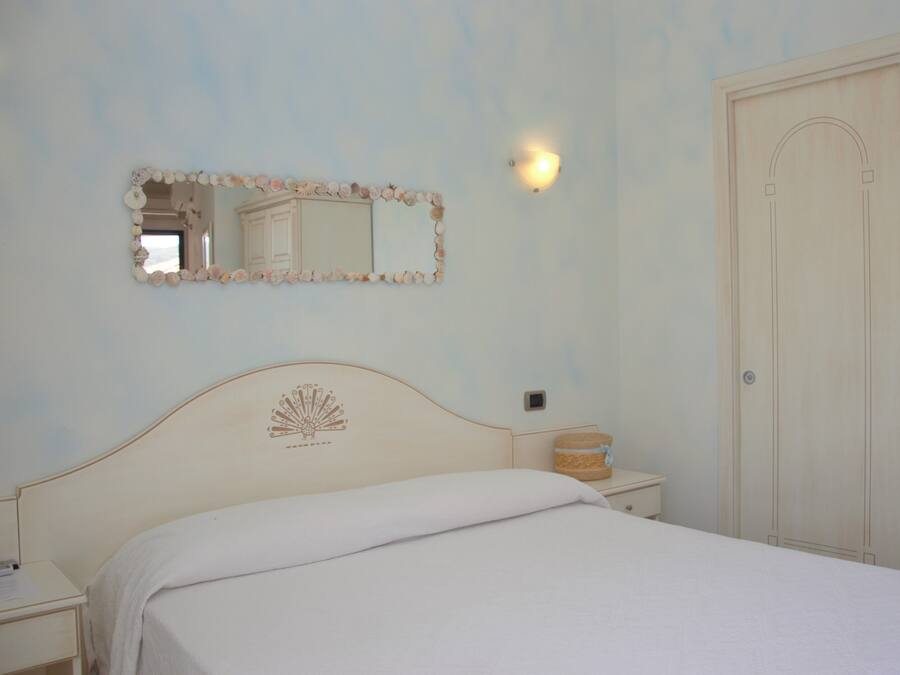 A bedroom with a bed, a nightstand, a mirror with a seashell frame, a wall-mounted light, and a door.