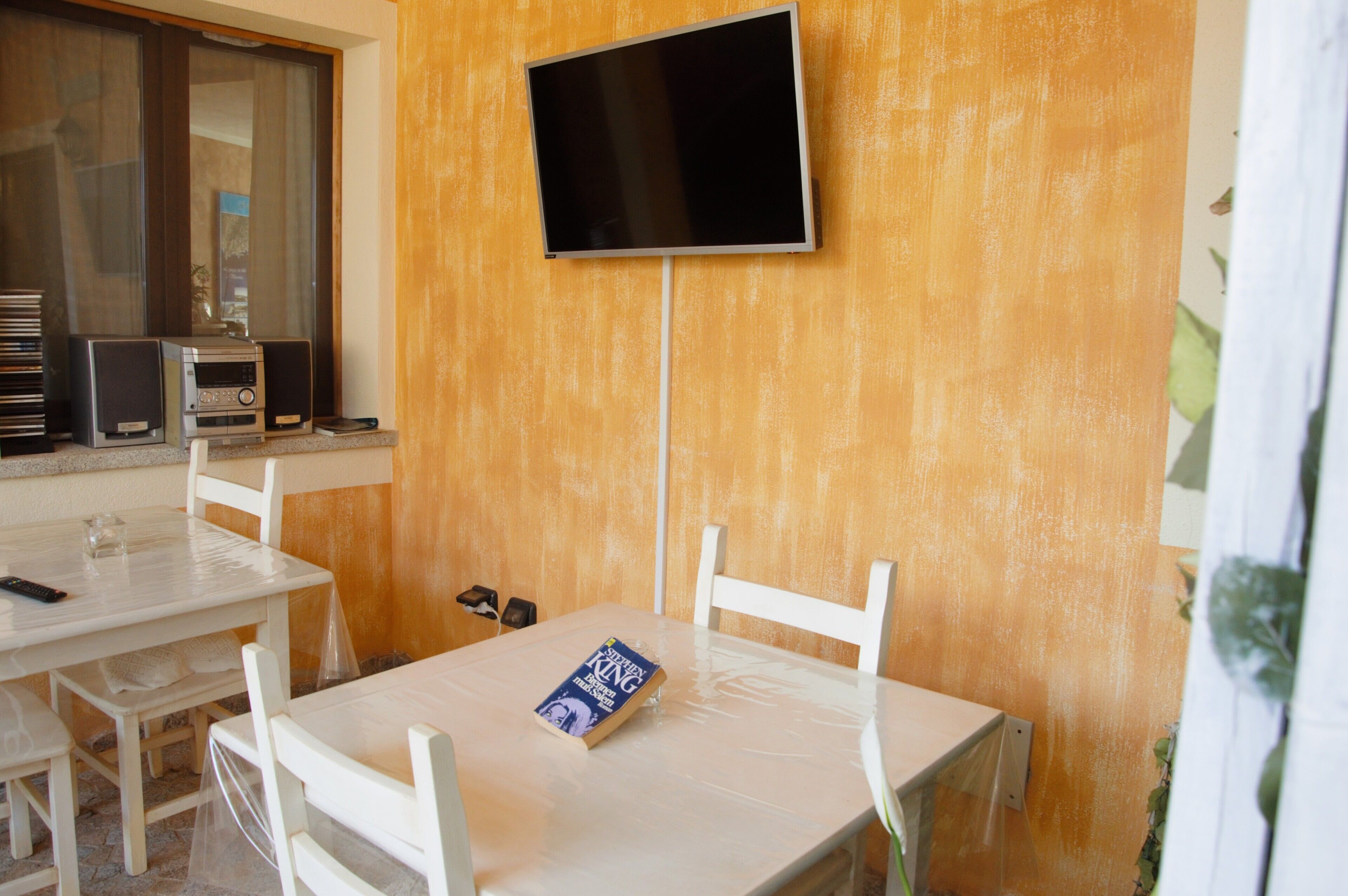 Ein Zimmer mit an der Wand montiertem Fernseher, einem Tisch mit Stühlen, einem Buch und Lautsprechern auf einem Regal.