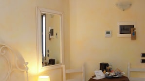 A hotel room with a bed, a bedside table with a lamp, a chair, a small table with a brown tablecloth, and an air conditioning unit on the wall.