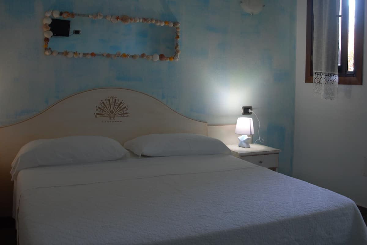 A bedroom with a bed, a nightstand with a lamp, a wall decoration, and a window with a curtain.