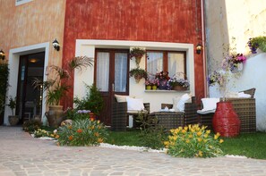 A building with a red facade, a door, and a window with hanging flower baskets. There is outdoor seating with cushions and a red vase. - Hotel Sa Cralla (Villasimius)