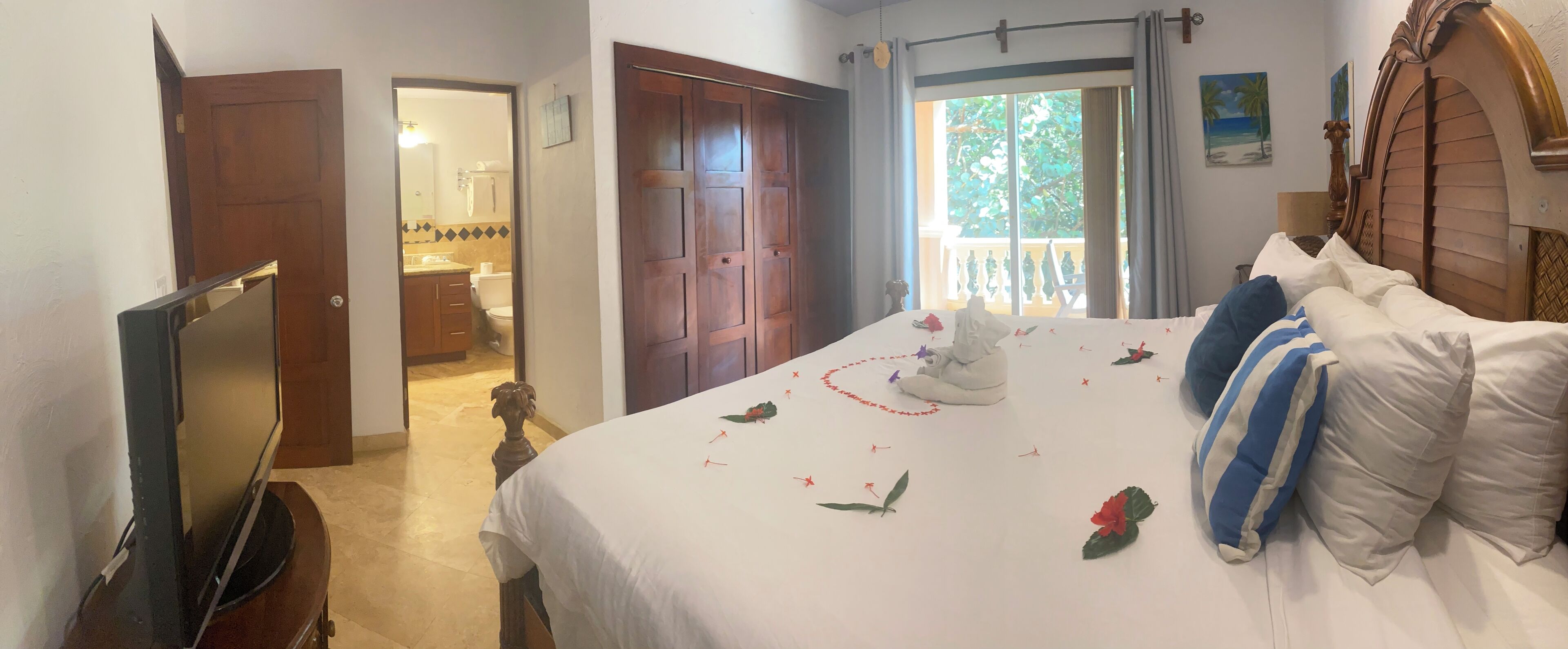 A hotel room with a bed, a TV, a balcony with a view of the outdoors, and a bathroom visible through an open door.