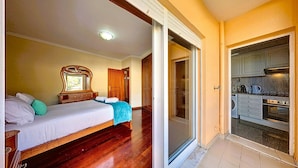 A bedroom with a bed, a dresser, and a mirror. There is a sliding glass door leading to a kitchen with a stove and cabinets. - CANIÇO CENTER APARTMENT (Santa Cruz)