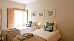 Habitación de hotel con dos camas, una mesita de noche con lámpara, una ventana con persianas y tres cuadros enmarcados en la pared.