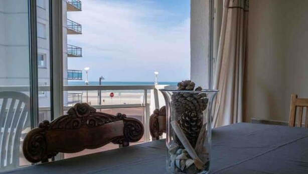 Ein Tisch mit einer klaren Vase, gefüllt mit Tannenzapfen und Muscheln, einem Automobil-Dekor im Vintage-Stil sowie Blick auf Strand und Meer durch große Fenster.