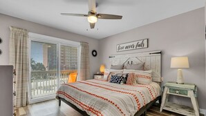 Un dormitorio con cama, una mesita de noche, ventilador de techo, ventana con cortinas y un cartel en la pared.