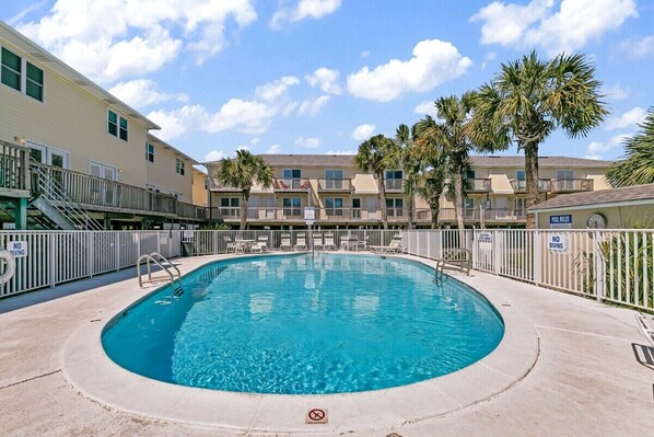 A swimming pool surrounded by a white fence, with palm trees and a multi-story building in the background.