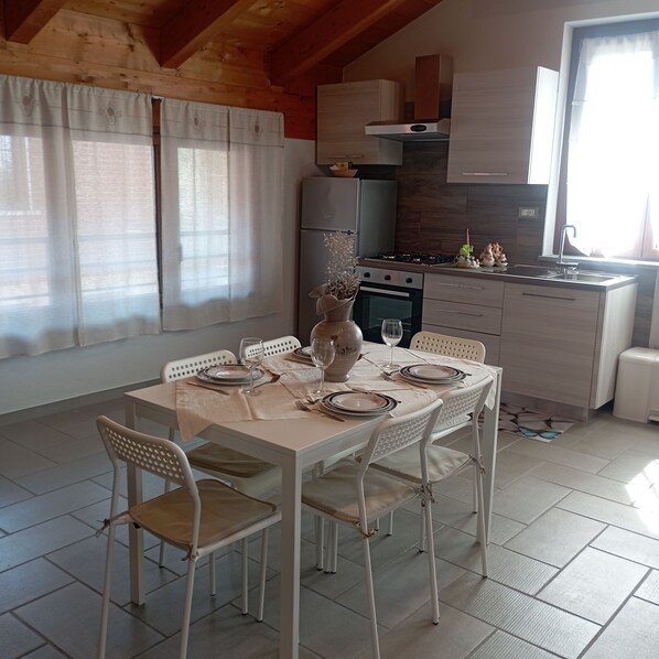 Un comedor con una mesa dispuesta para cuatro personas, sillas blancas, una cocina al fondo y una ventana con cortinas.