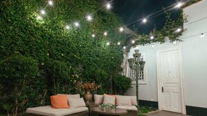 A cozy outdoor seating area with a wicker sofa and a round table, surrounded by lush greenery and string lights.