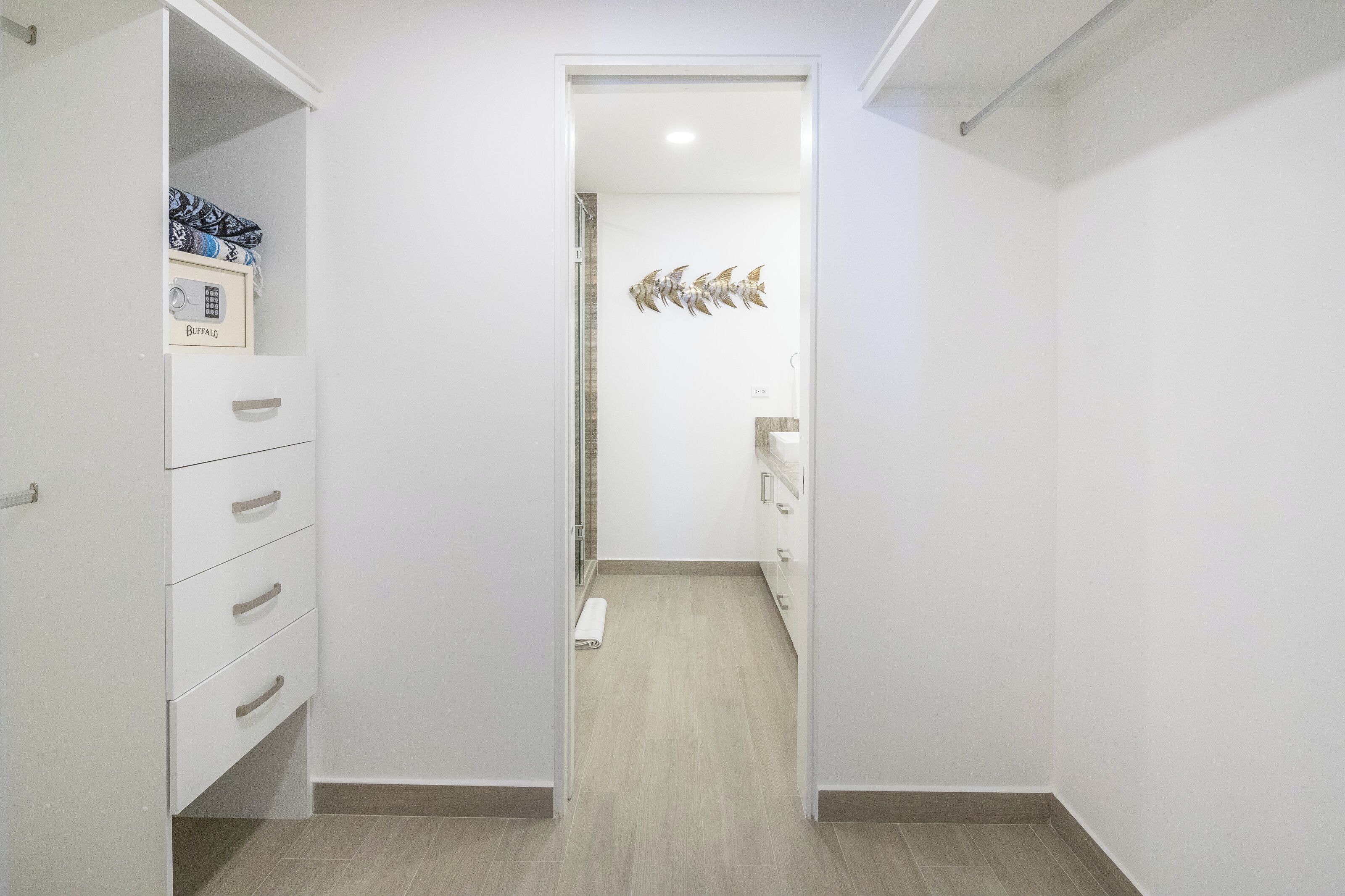 Ein schmaler Flur mit einem weißen Kleiderschrank links, einem Duschbereich mit einer gefliesten Wand und einem Handtuch auf dem Boden sowie einem kleinen Waschbecken rechts.