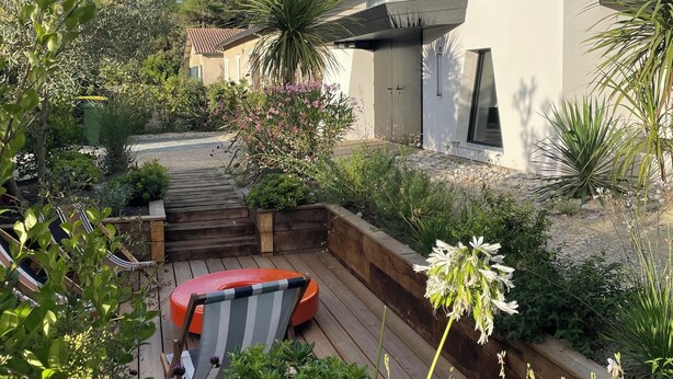 Une maison moderne dotée d’une terrasse en bois, d’une chaise rouge et de diverses plantes.