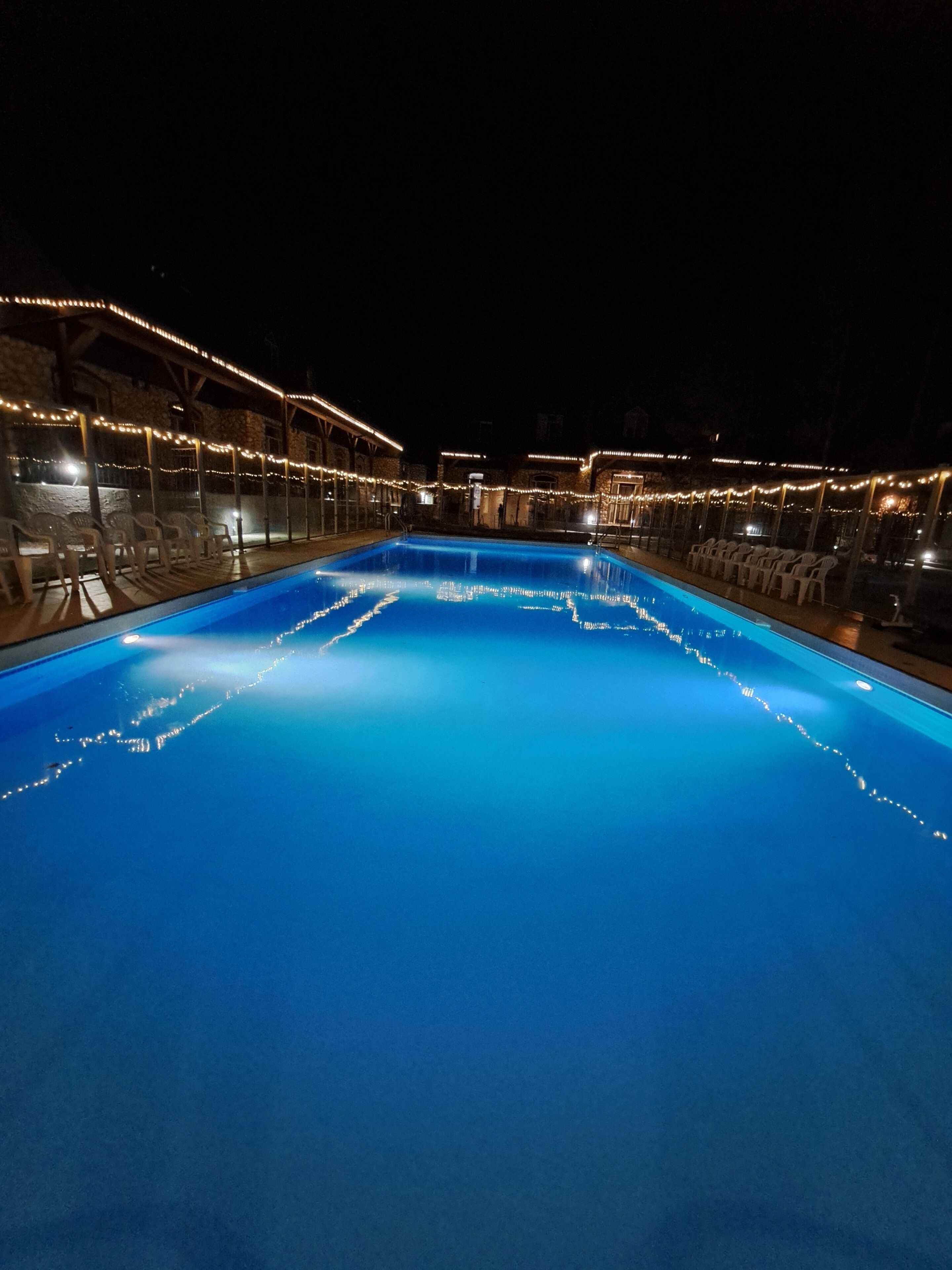 A swimming pool with a clear blue water surface, surrounded by white chairs and string lights.