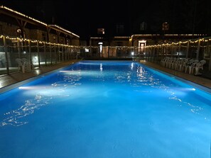 En swimmingpool med ett klart, blått vatten, omgiven av en byggnad i sten med ett trädäck och ljusslingor.