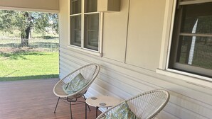 Un porche abrité avec deux fauteuils en osier et une petite table, des murs extérieurs blancs et une fenêtre donnant sur un espace verdoyant.