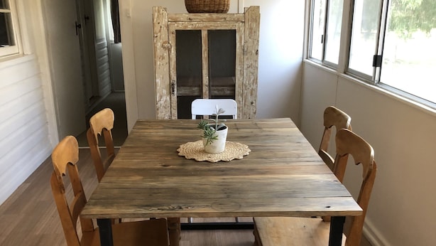 Une table de salle à manger en bois, accompagnée de six chaises, d’un banc blanc et d’une petite plante en pot posée sur un set de table en fibres tressées.