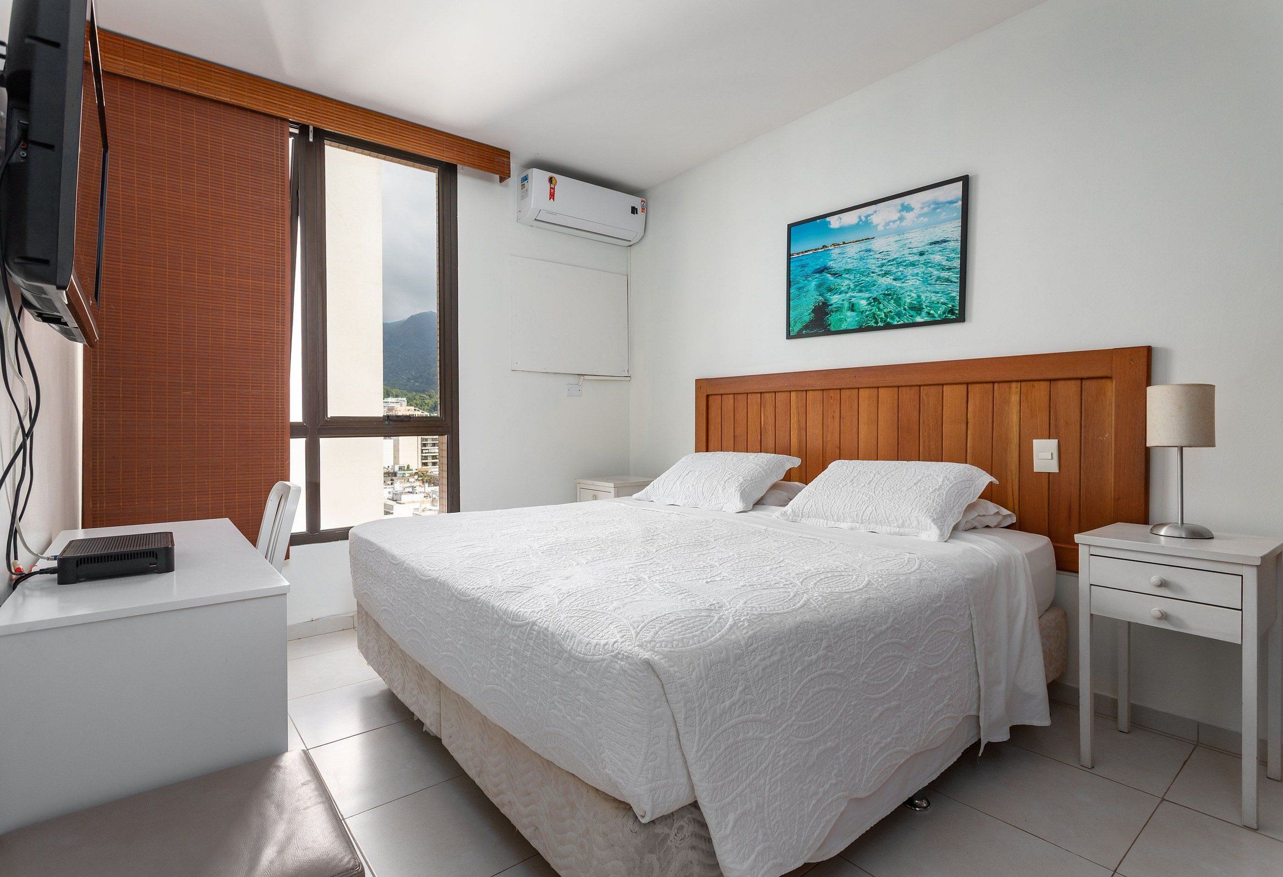 Une chambre d’hôtel comprenant un lit, un bureau, une chaise, une table de chevet, une lampe et une fenêtre avec des stores.