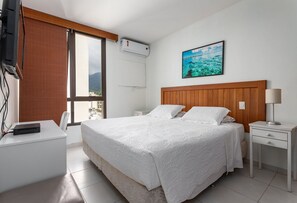 Habitación de hotel con cama, escritorio, silla, mesita de noche, lámpara y ventana con persianas.