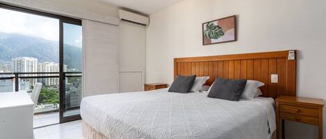 A hotel room with a large bed, a wooden headboard, a bedside table, a nightstand, a window with a view of buildings and mountains, and a painting on the wall.