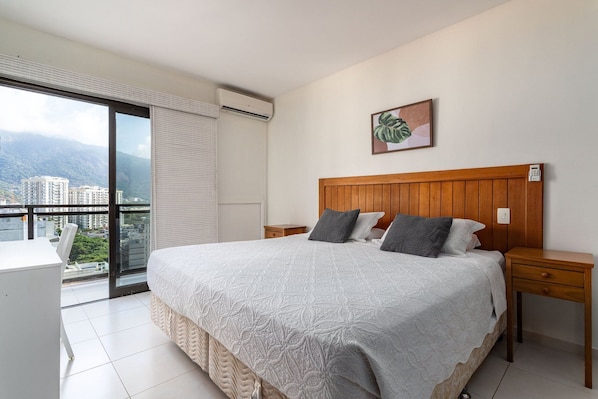 A hotel room with a large bed, a wooden headboard, a bedside table, a nightstand, a window with a view of buildings and mountains, and a painting on the wall.