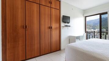 Un dormitorio con un armario amplio, una cama con edredón blanco, un televisor montado en la pared y vistas a un paisaje urbano a través de una ventana.