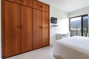 A bedroom with a large wardrobe, a bed with a white duvet, a television mounted on the wall, and a view of a cityscape through a window.