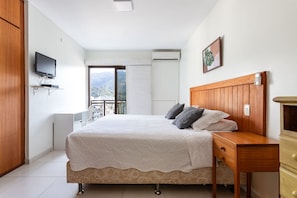A bedroom with a bed, a TV mounted on the wall, a window with a view of buildings and mountains, and a wooden nightstand. - (1306) Apartamento 2 Suítes e Vista para o Cristo (Rio de Janeiro)