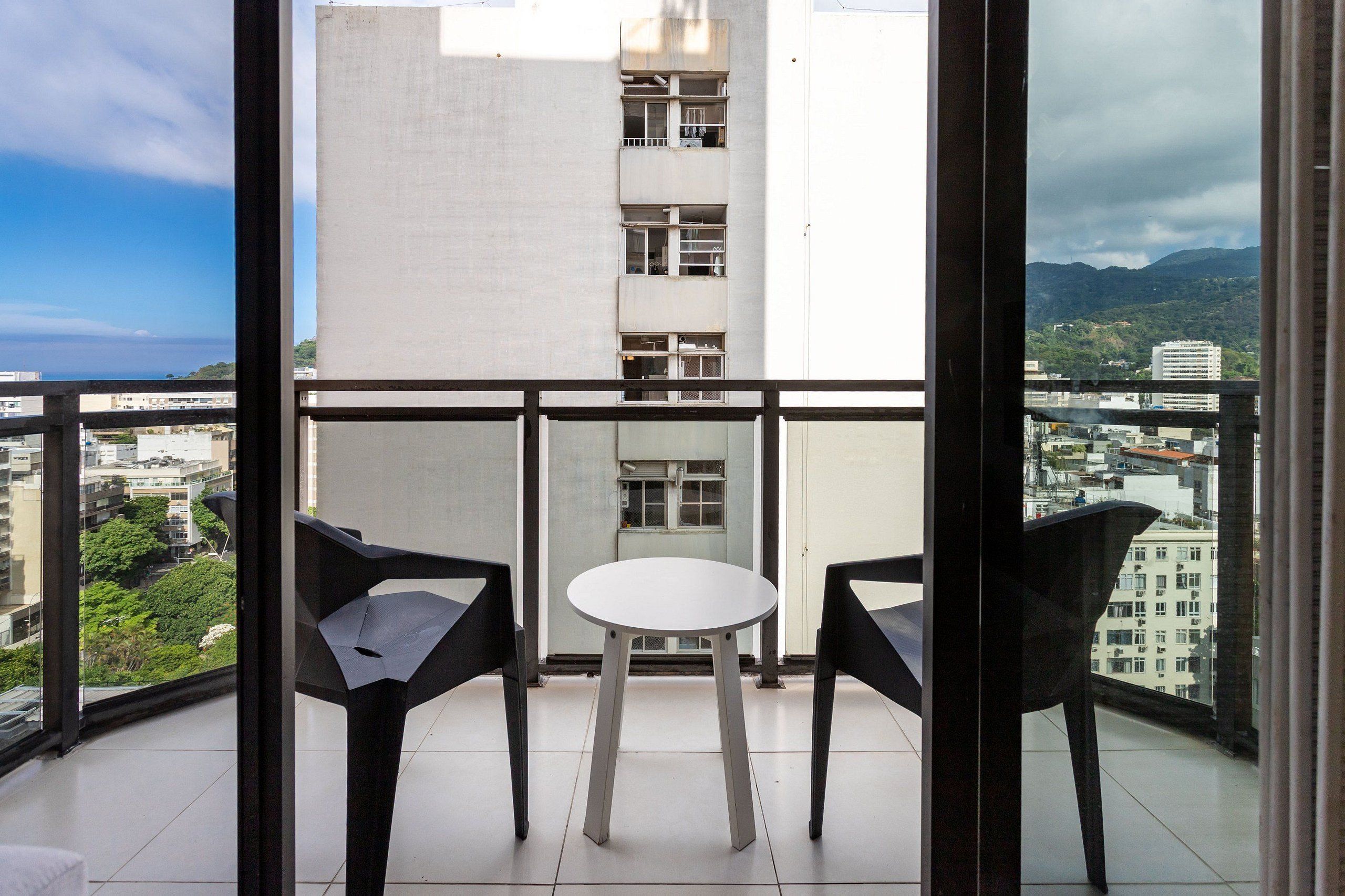 A balcony with two chairs and a small table, overlooking a cityscape with buildings and greenery.