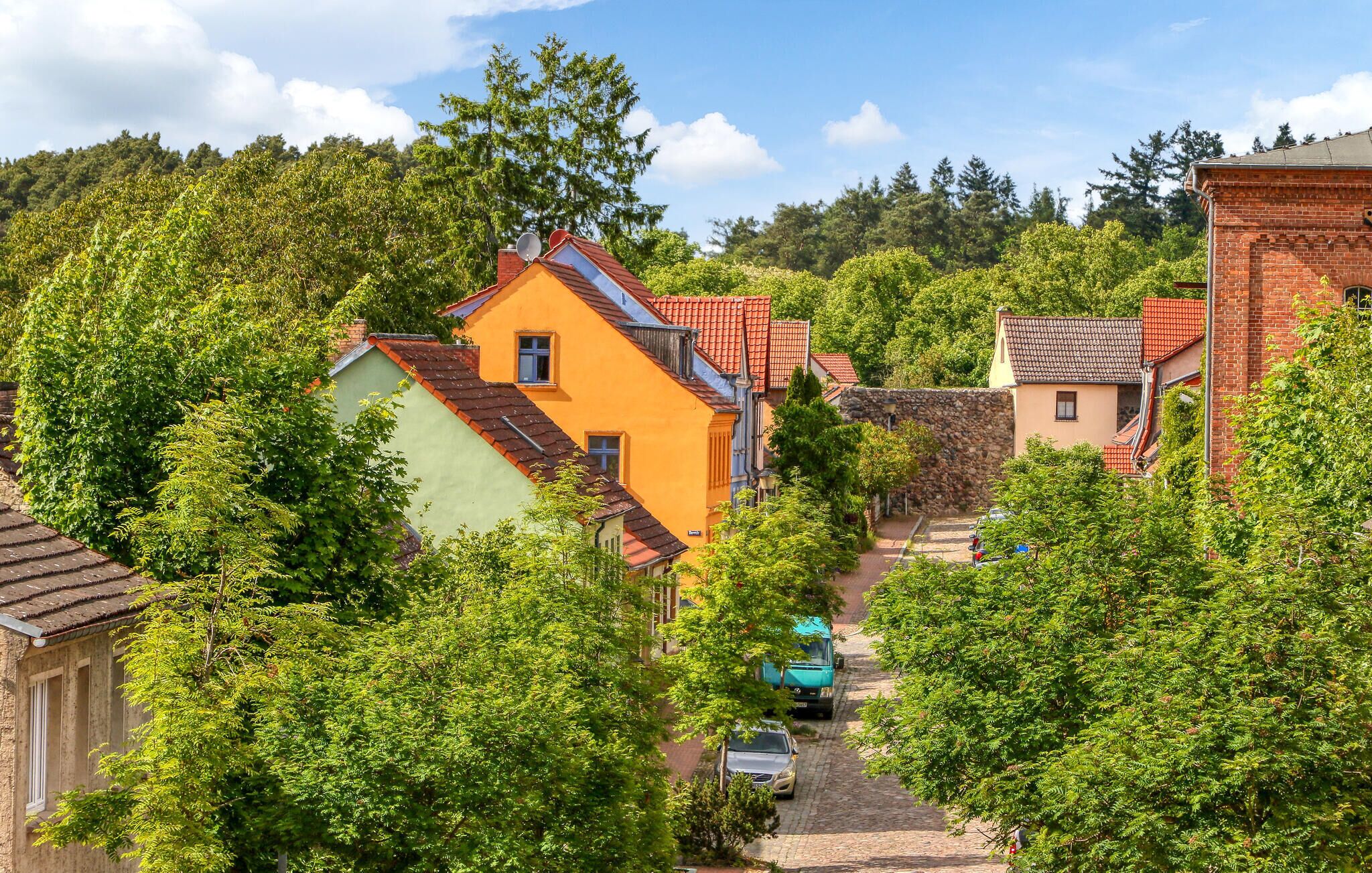 Une ruelle étroite pavée, bordée de maisons aux couleurs vives et entourée d’arbres verdoyants.