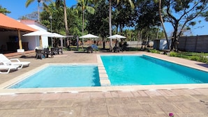 Ein rechteckiger Schwimmteich mit zwei Bereichen, umgeben von einer gefliesten Terrasse. Weiße Liegestühle und Sonnenschirme sind vorhanden, sowie ein überdachter Sitzbereich mit Tischen und Stühlen.