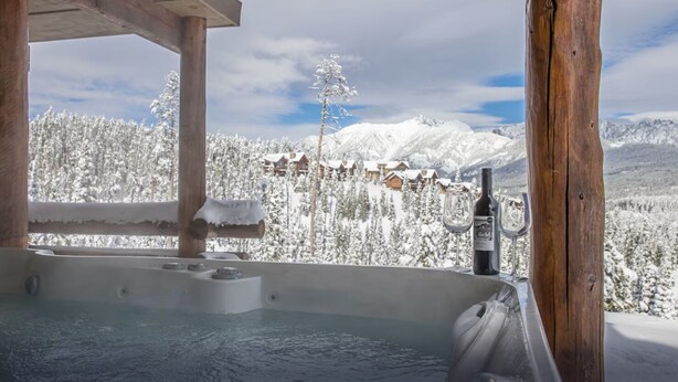 Ett bubbelbad med en träkonstruktion, en flaska vin och glas på ett bord, med utsikt över ett snöklätt berglandskap med stugor.