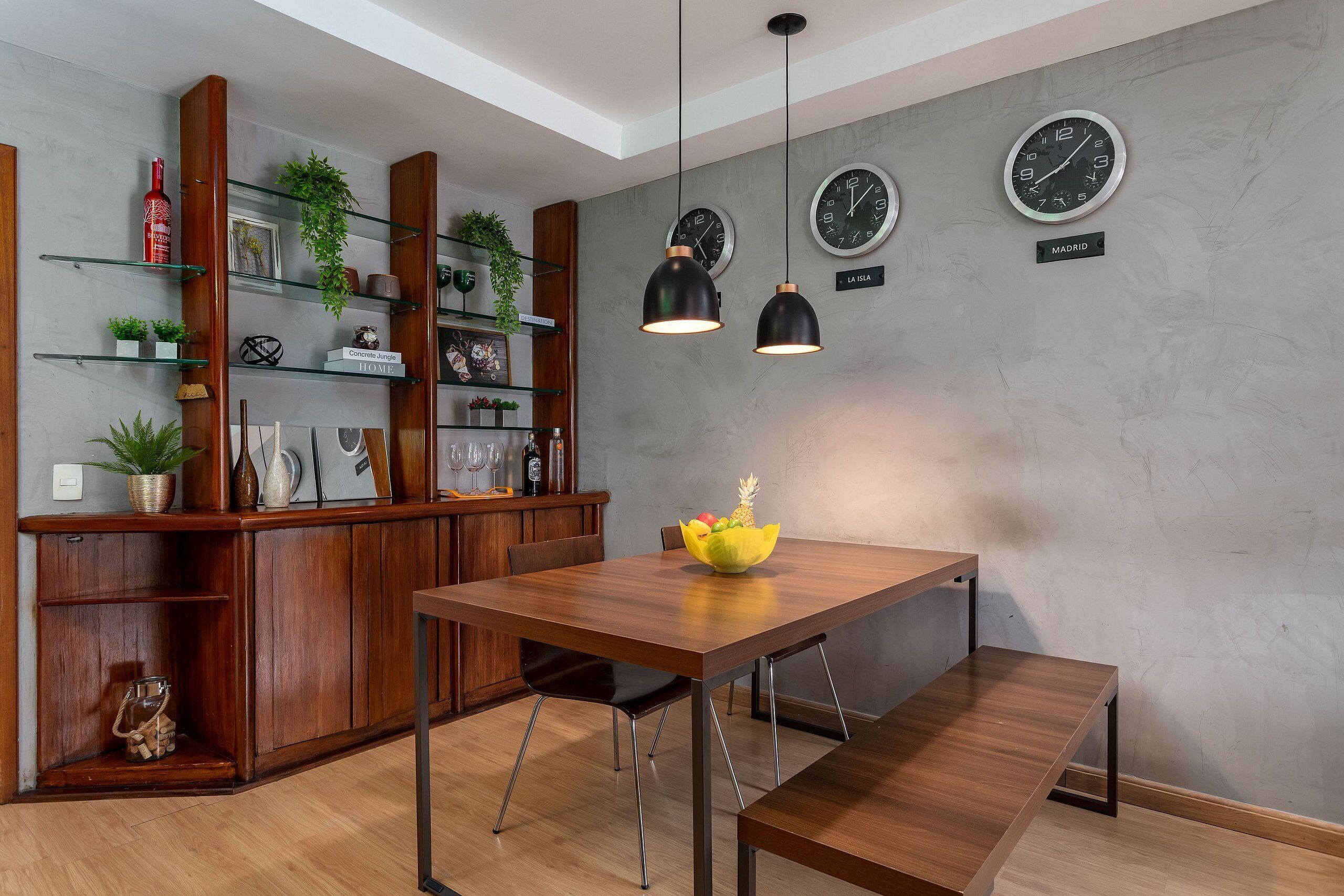 A modern dining area with a wooden table, bench, and pendant lights. There are two clocks on the wall and a shelf with decorative items.