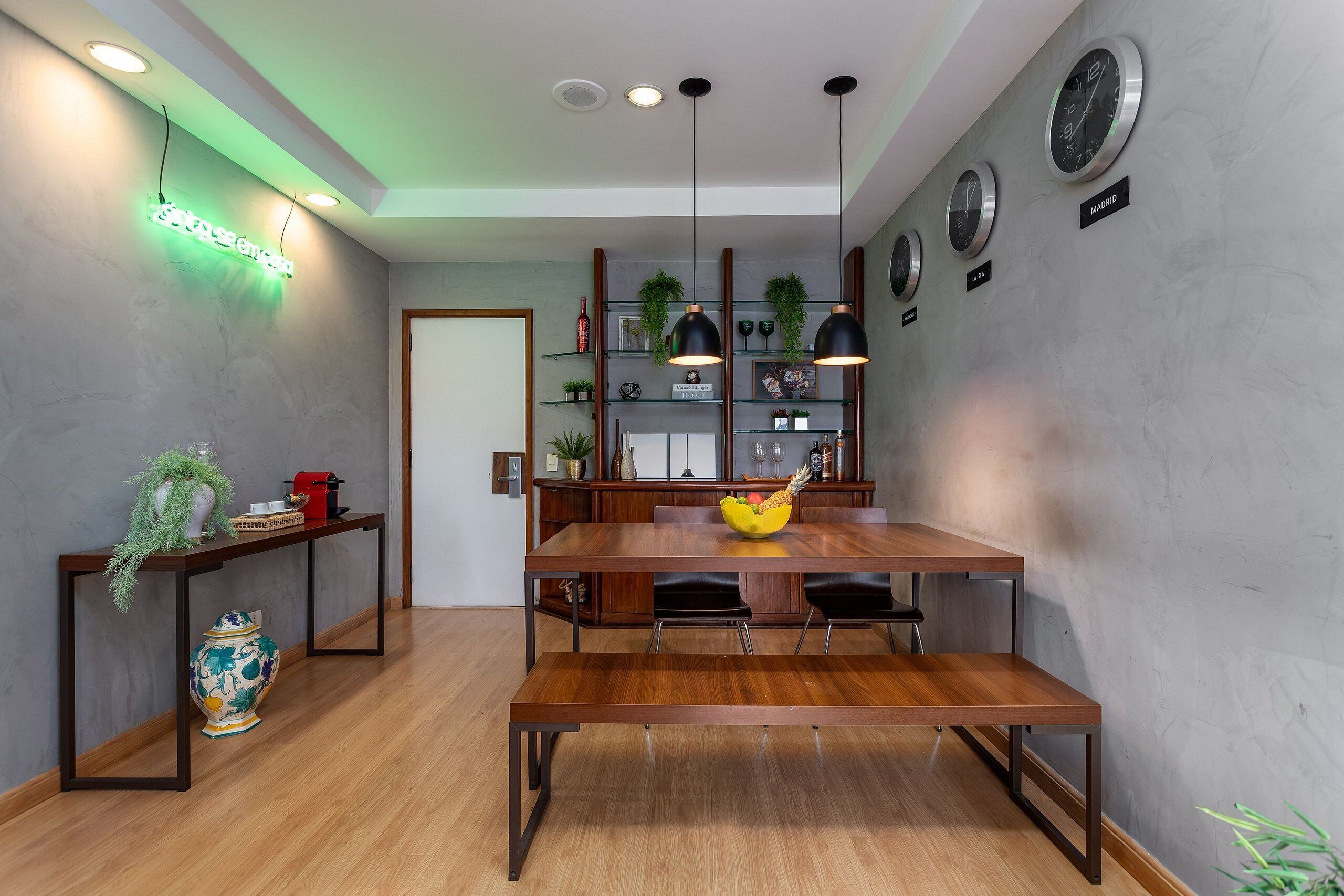 Un comedor con mesa y bancos de madera, una estantería con adornos y relojes de pared.