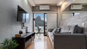 Una sala de estar moderna con un sofá gris, un televisor de pantalla plana montado en la pared, un mueble de entretenimiento negro y un balcón con vistas a la montaña.