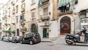 Une rangée de voitures garées le long d’une rue pavée, dans un quartier historique de la ville, bordée d’immeubles à plusieurs étages, dont certains sont dotés de balcons et de plantes d’extérieur.