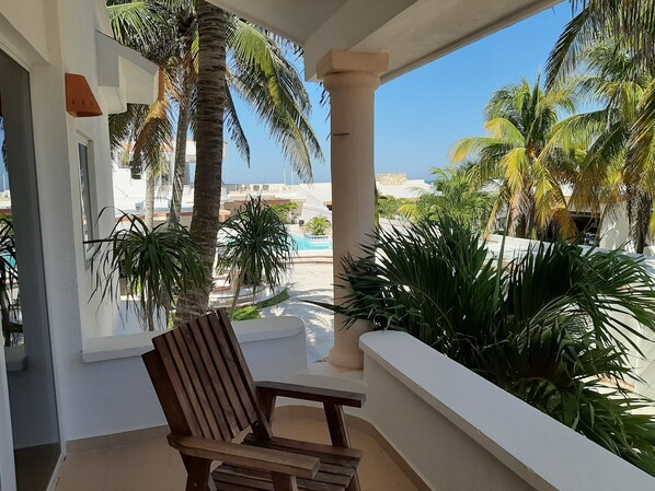 Un balcon avec une chaise en bois, des palmiers et une vue sur une piscine et des immeubles.