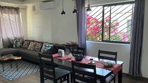 A living room with a sofa, dining table, chairs, and a window with a view of pink flowers.