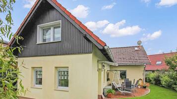 Een huis met twee verdiepingen, voorzien van een rode pannendak en een klein terras met zitplaatsen.