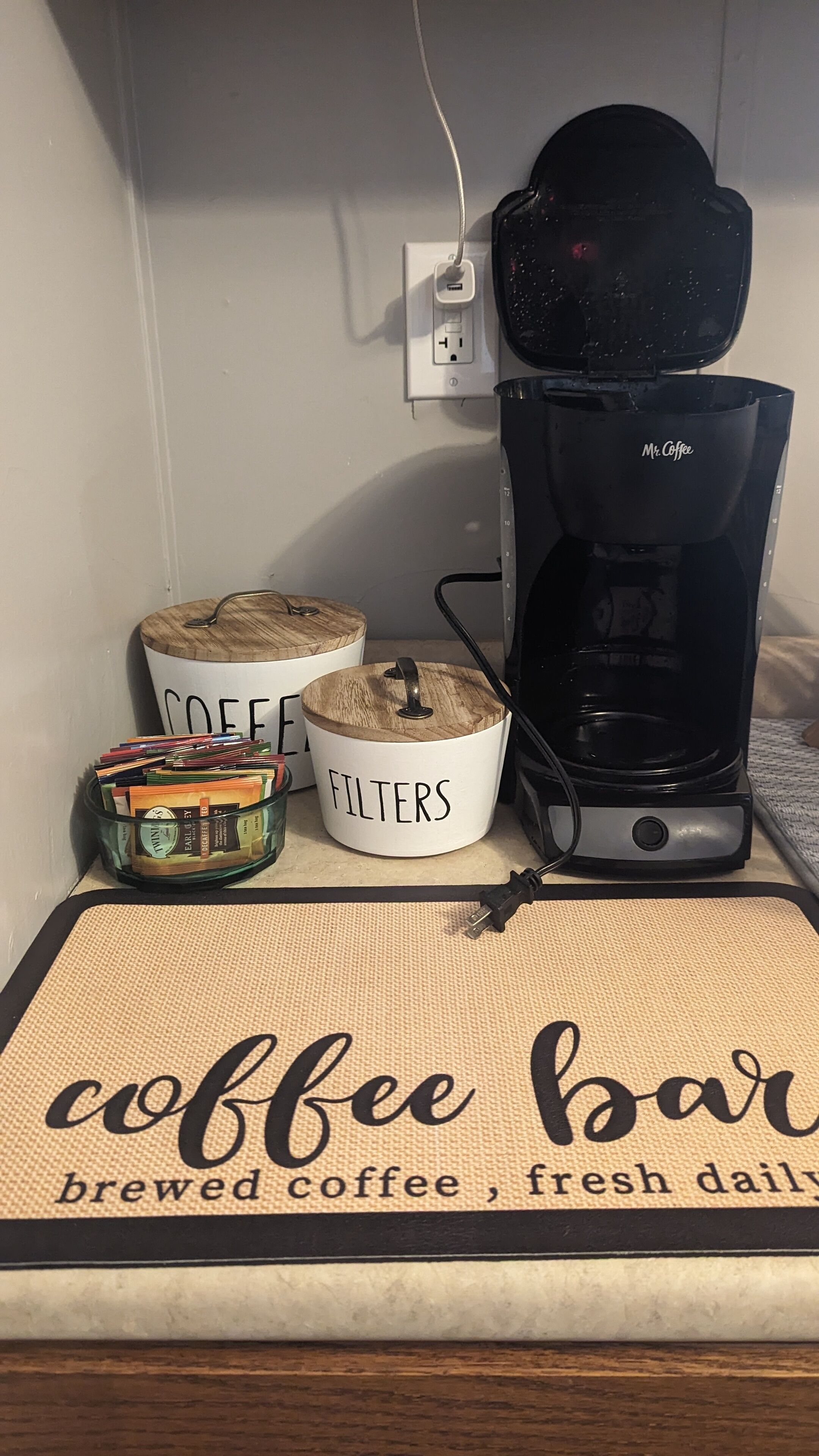 En kaffestation med en kaffebryggare, två märkta behållare för kaffe och filter, samt en matta med texten ”nybryggt kaffe från vårt kaffebord, serveras varje dag”.