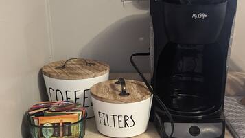 En kaffestation med en kaffebryggare, två märkta behållare för kaffe och filter, samt en matta med texten ”nybryggt kaffe från vårt kaffebord, serveras varje dag”.
