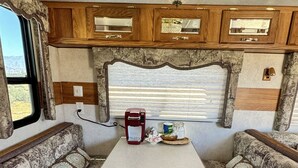 Interior compacto de una caravana con kitchenette, dos áreas de estar con asientos acolchados, cafetera y lámpara de araña.