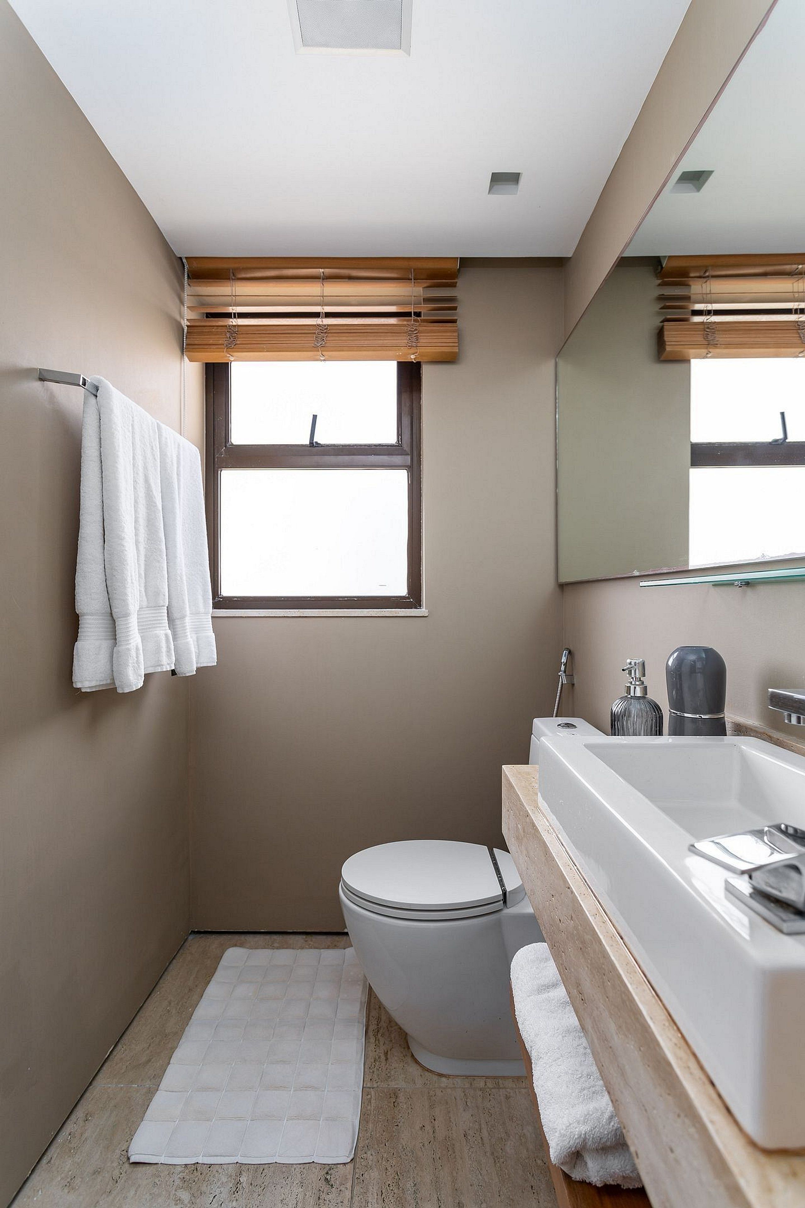 A modern bathroom with a toilet, a bathtub, and a shower. There is a window with blinds, a towel rack, and a mirror above the sink.