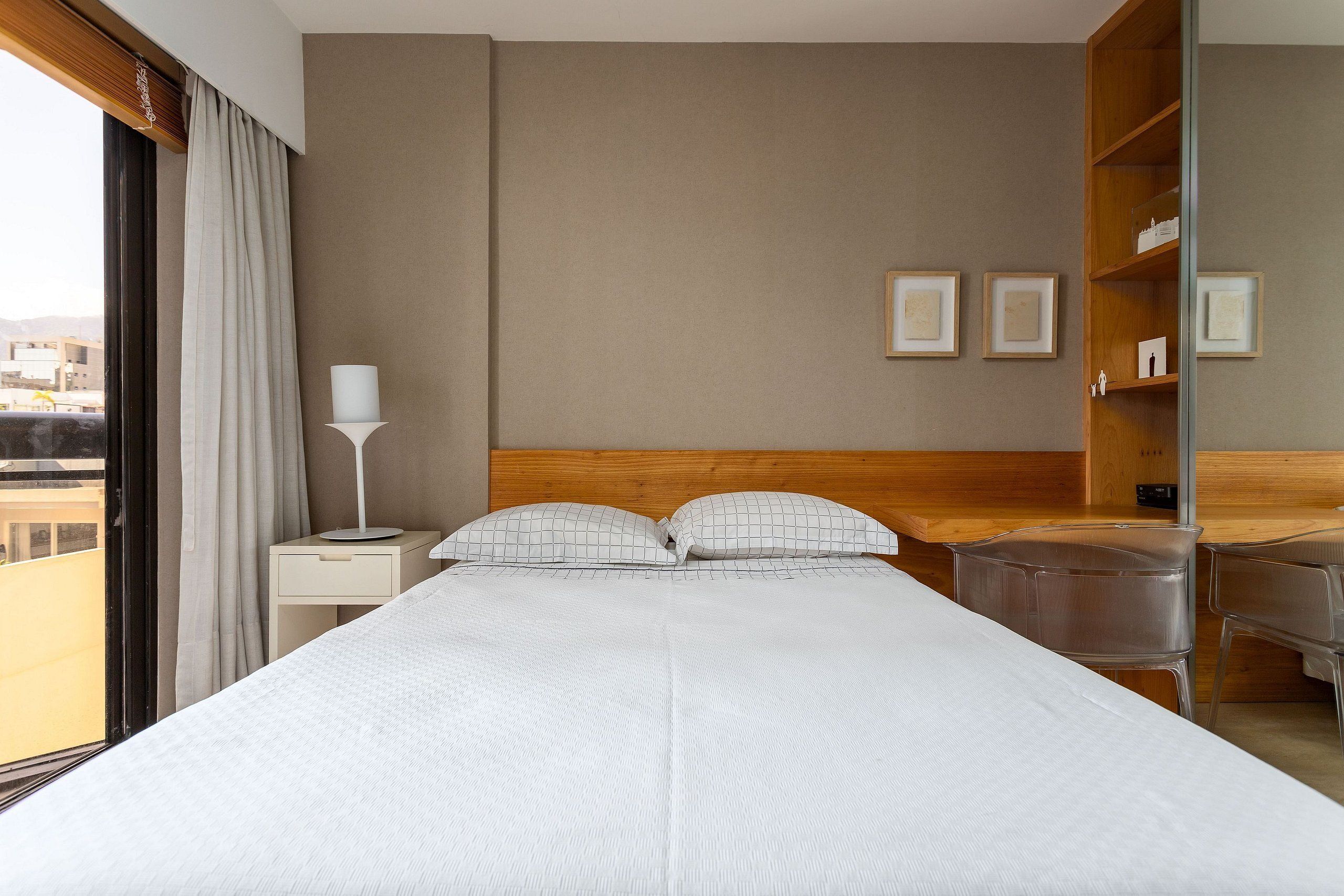 A hotel room with a large bed, a bedside table with a lamp, a wooden headboard, and a view of the city.