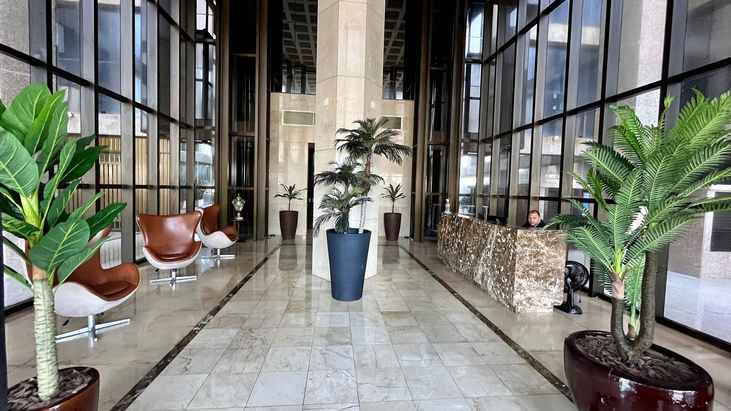 Un vestíbulo moderno con piso de mármol, ventanas amplias y varias áreas de estar con sillas y plantas en macetas.